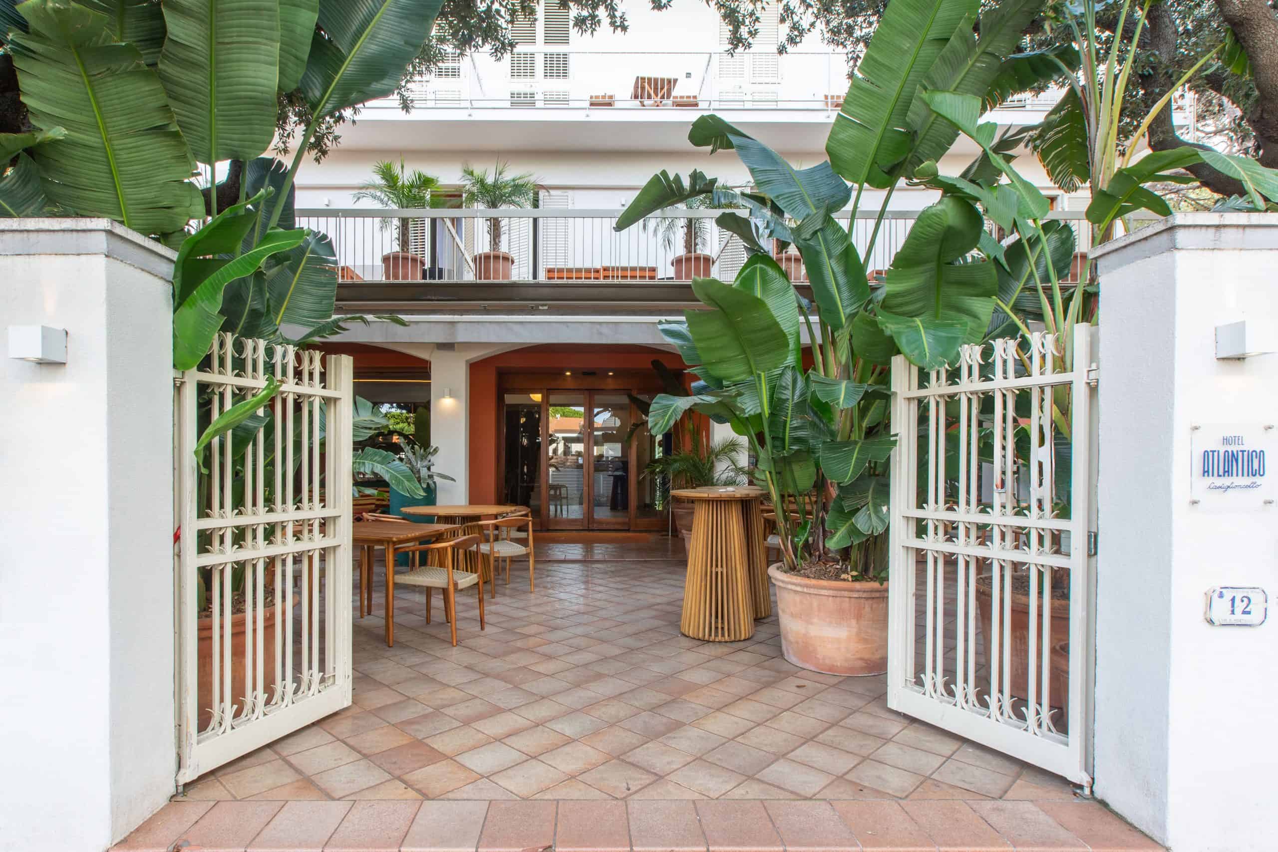 Vista dell'ingresso dell'Hotel Atlantico a Castiglioncello, con un elegante cancello bianco aperto che conduce a un patio accogliente con tavoli in legno e piante rigogliose.